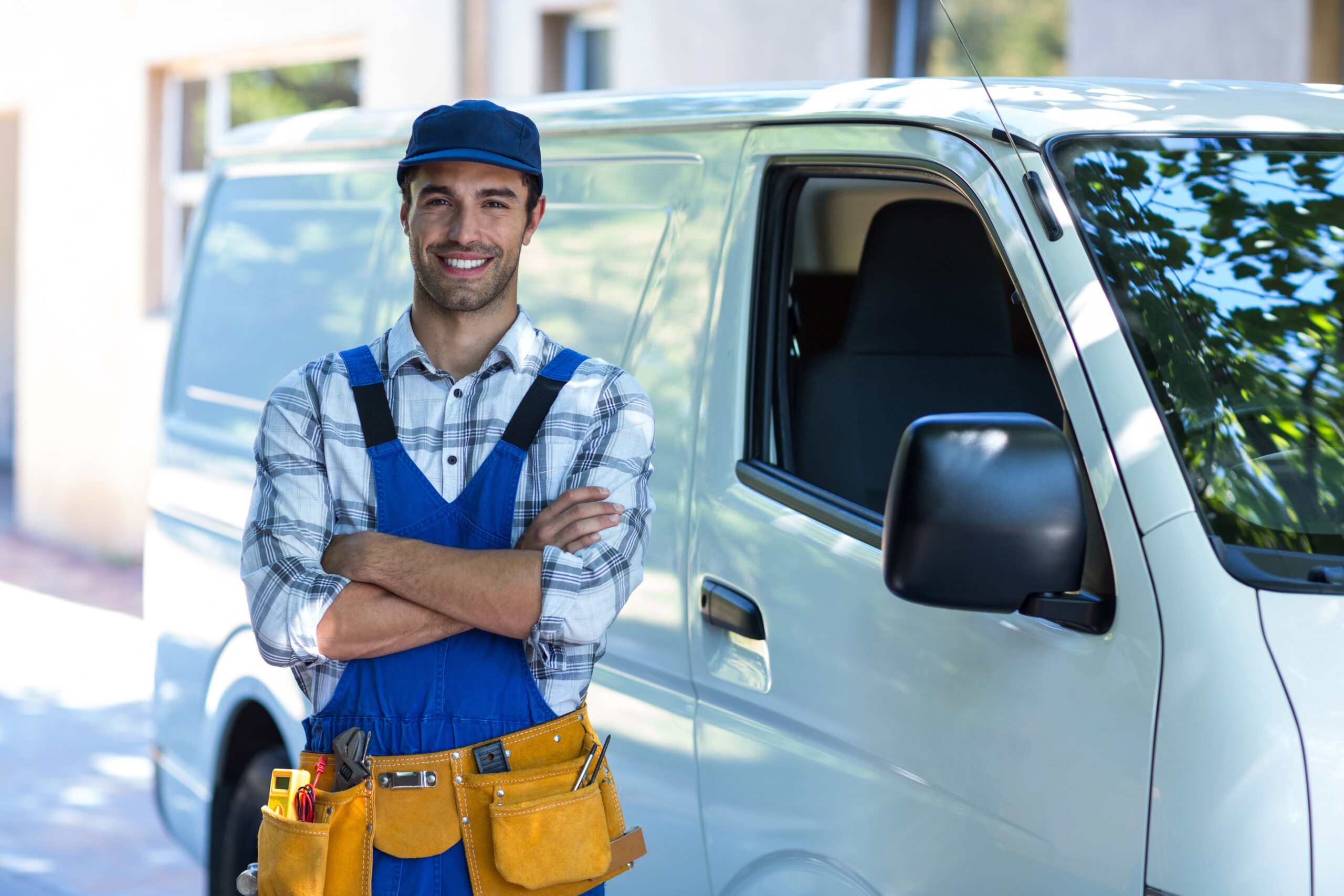 Maintenance technician in a yellow work belt and blue hat stands in front of his white work van with arms crossed