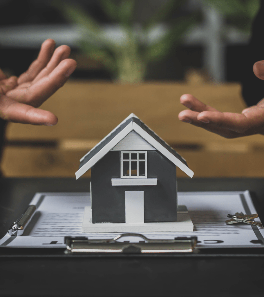 A small grey wooden house on clipboard with papers, and two human hands around it.
