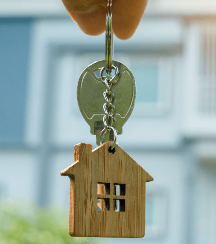 Single key with a small wooden cutout home keychain, being dangled by the keyring between two fingers.