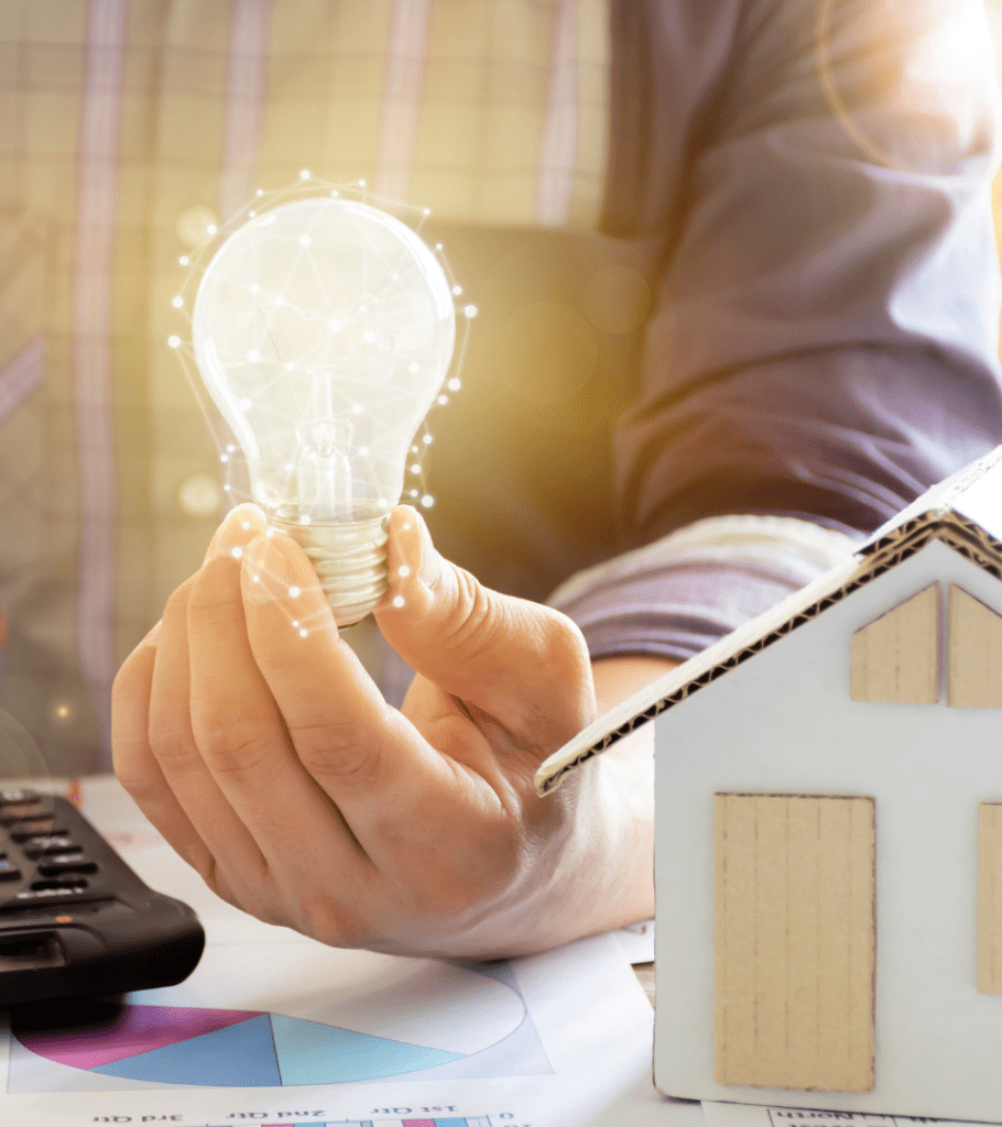 A man holds an illuminated lightbulb over a calculator, cardboard house cutout, and an open spreadsheet, representing getting ideas to save money on operating costs for investment properties.