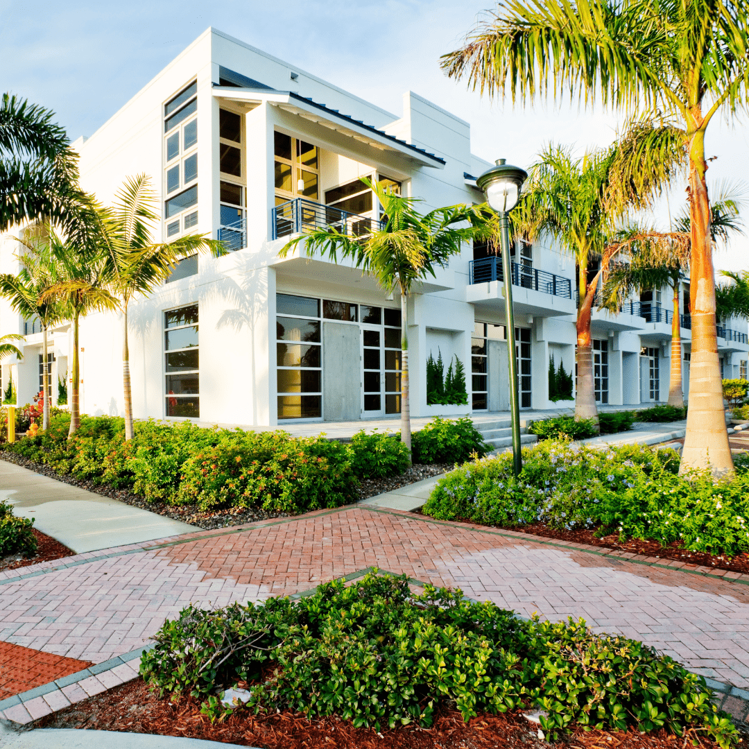 An exterior view of a white, rectangular industrial building with rows of large, reflective windows across the facade. Several palm trees in the front lawn provide landscaping. The image shows the building on a sunny day with clear skies and a stone pathway to the entrance.