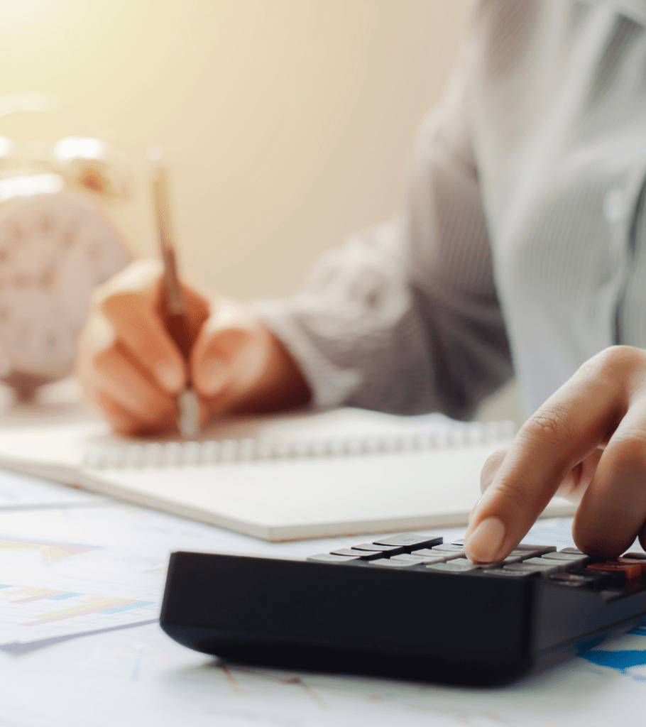 A man sits at a desk, as he works on his taxes. In one hand he holds a pen, hovering over an open notepad filled with calculations. His other hand types numbers into a calculator. Behind him, an alarm clock, indicating he has been working to complete his tedious financial paperwork before the tax deadline.