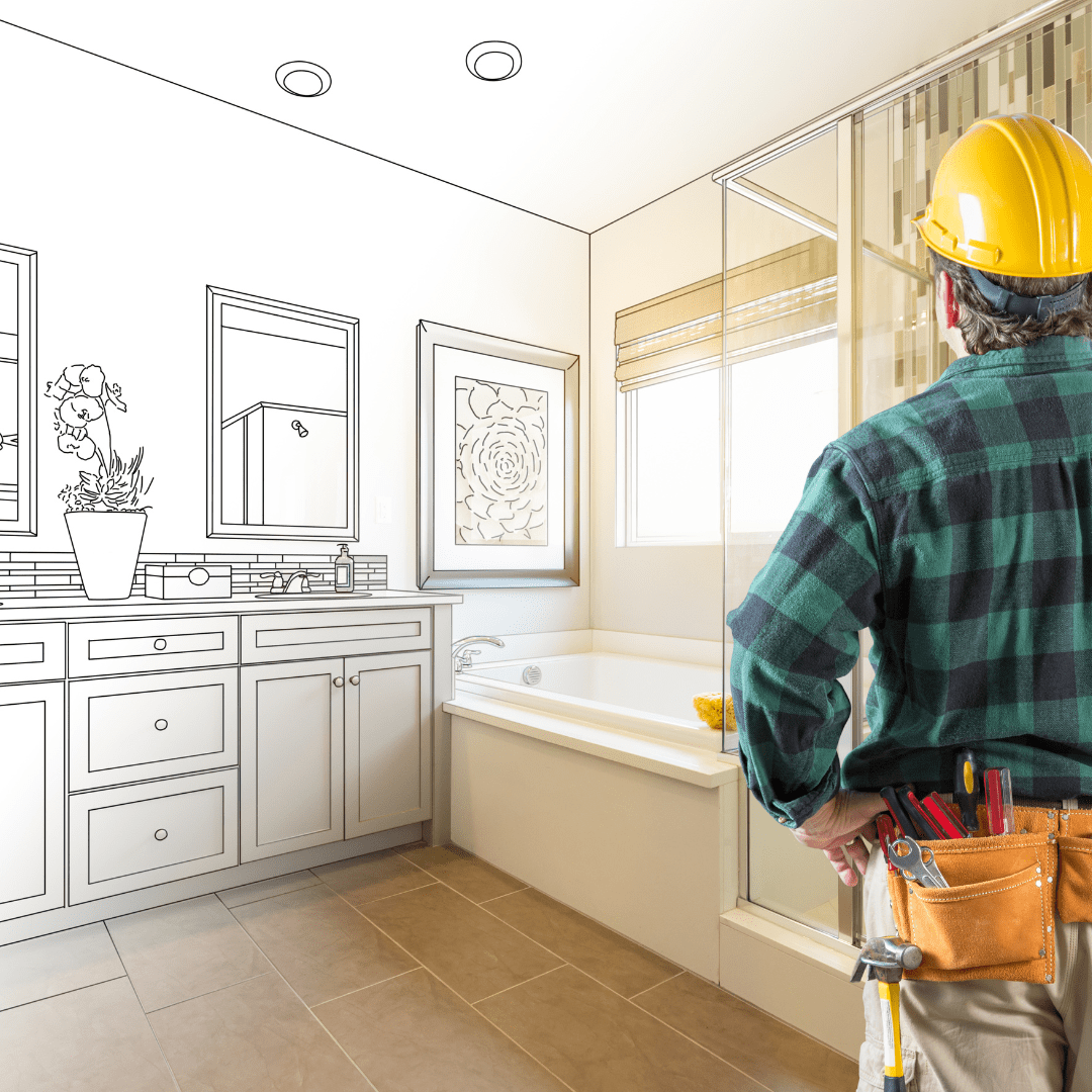 A man wearing a yellow hard hat and tool belt looks pensively at blueprints for a bathroom renovation in progress. The left side of the image is black and white showing the unfinished bathroom, while the right side fades into full color showing the stylish finished bathroom design.