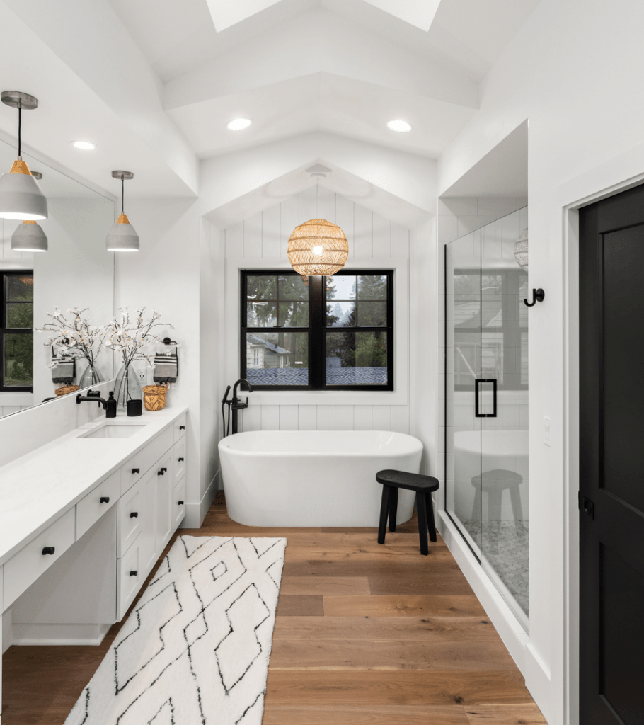A bright white wall-length vanity sits atop wood floors in this bathroom featuring a free standing tub positioned below a pane window for natural light, an adjacent glass door shower, and a round fixture hanging above the tub.