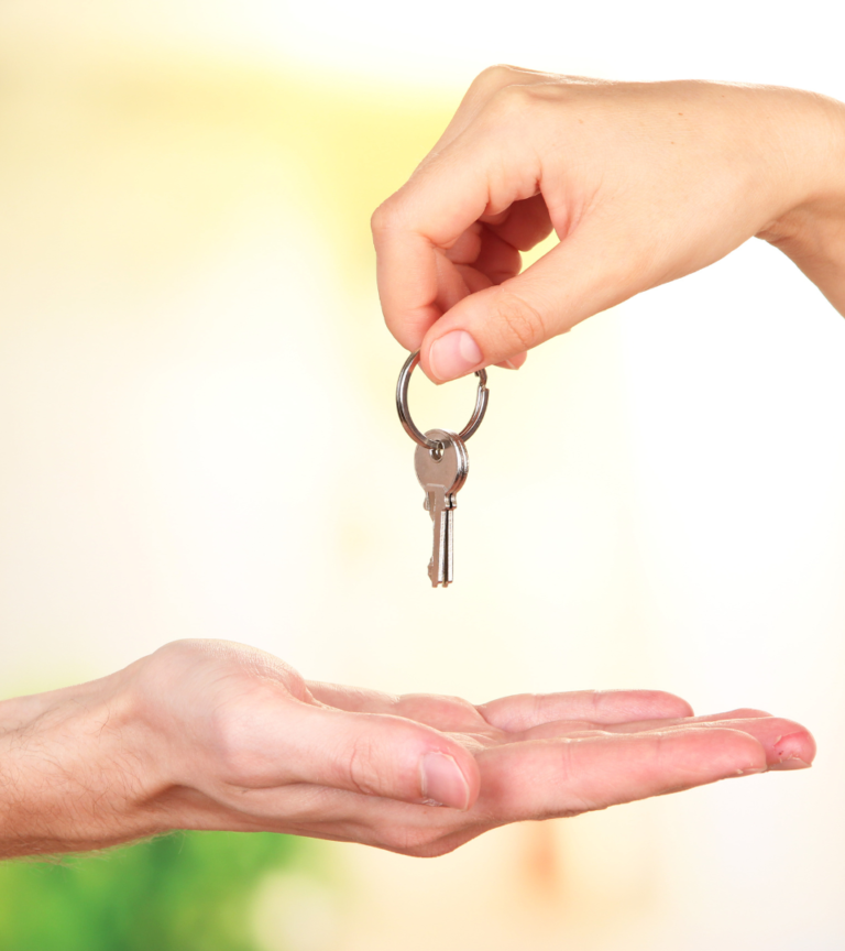 Two hands, one handing over a key to the other outstretched hand, implying the turnover or transfer of a rental property to a new tenant.